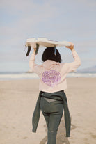 A person strolls along the sandy beach, surfboard in hand, showcasing a pink Yin-Yang Crop Hoodie from the Spring 2024 collection paired with black wetsuit bottoms.