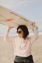 Individual standing on a sandy beach, holding a surfboard overhead while wearing sunglasses and the 80s-inspired Yin-Yang Crop Hoodie from the Spring 2024 collection.