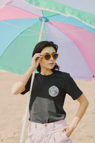 A woman relaxes beneath a vibrant beach umbrella, wearing sunglasses and a Vintage Black Yin-Yang Tee from the 80s-inspired collection, ideal for a sunny day at the beach.