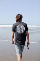 Walking along the beach, a person in an 80s-inspired Yin-Yang Tee - Vintage Black and shorts holds their sunglasses, basking in the sun.