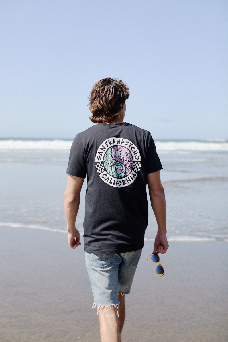 Walking along the beach, a person in an 80s-inspired Yin-Yang Tee - Vintage Black and shorts holds their sunglasses, basking in the sun.