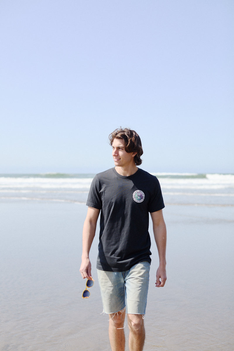 A man in a Vintage Black Yin-Yang Tee and denim shorts strolls along the beach, holding sunglasses as waves crash gently in the background.