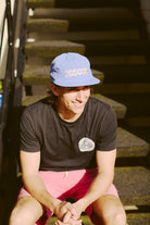 With a beaming smile, a person proudly dons a Checked Out Hat made of blue nylon from the Spring 2024 collection and pairs it with a black t-shirt while sitting on outdoor steps, basking in the sunlight.