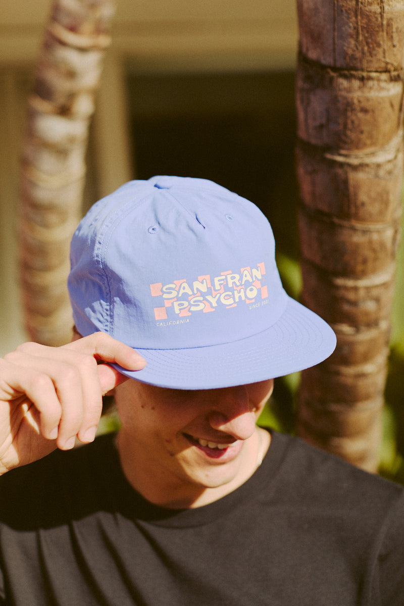 A person in a black shirt sports the Checked Out Hat from San Fran Psycho's Spring 2024 collection under a palm tree, channeling an 80s skate-inspired vibe.