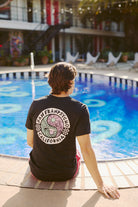 A person sits by the pool in a San Franpsycho Yin-Yang Tee - Vintage Black made from soft Airlume cotton, with string lights twinkling in the background.