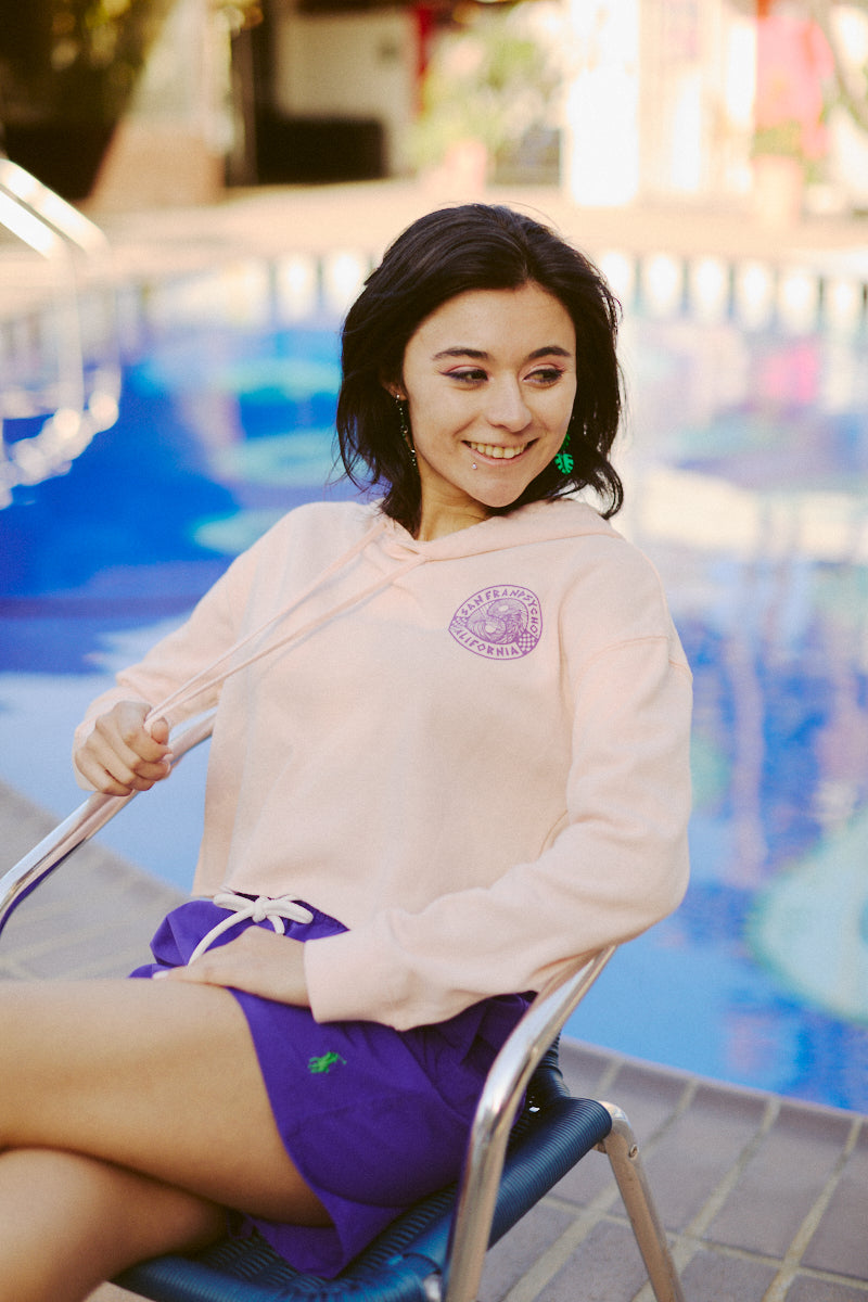 A person in an 80s-inspired hoodie sits on a chair by the pool, smiling and holding the strings of their Yin-Yang Crop Hoodie from the Spring 2024 collection.