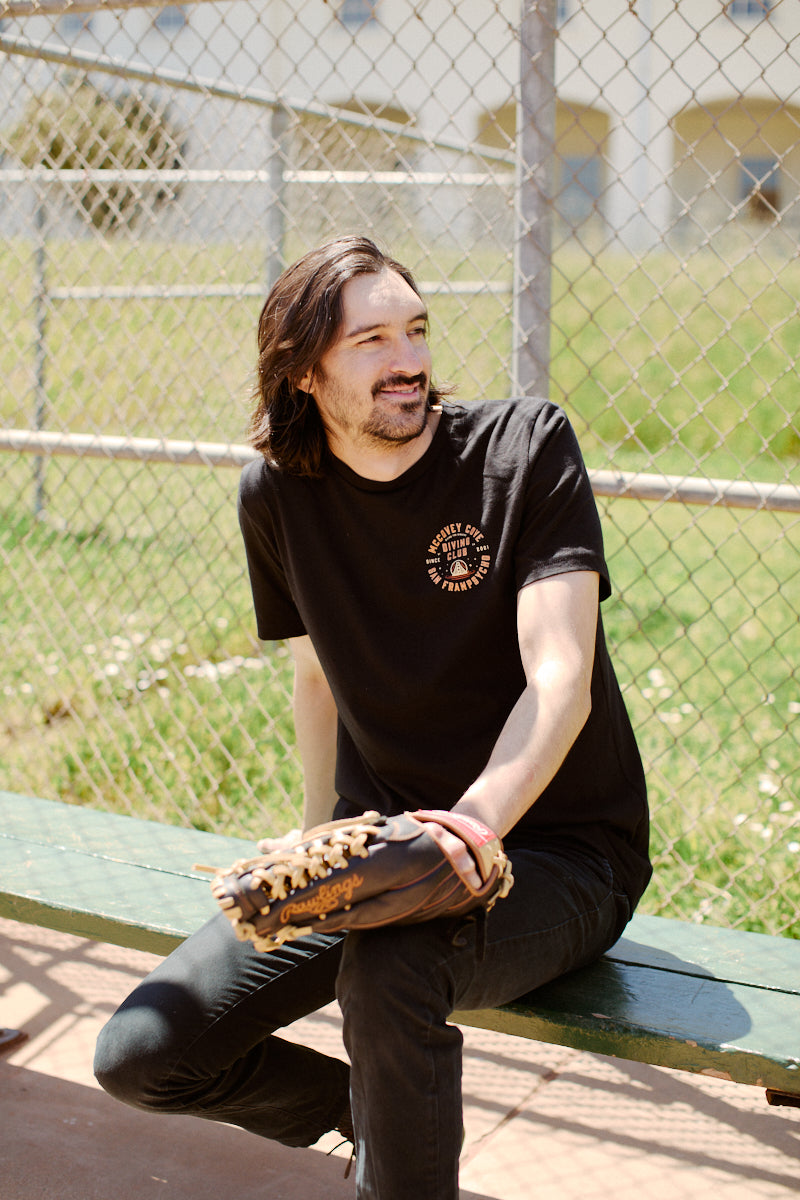 A man sits on a bench with a baseball glove, donning Giants gear—a McCovey Cove Diving Club Tee made from 100% cotton—in a sunny outdoor setting near a chain-link fence.