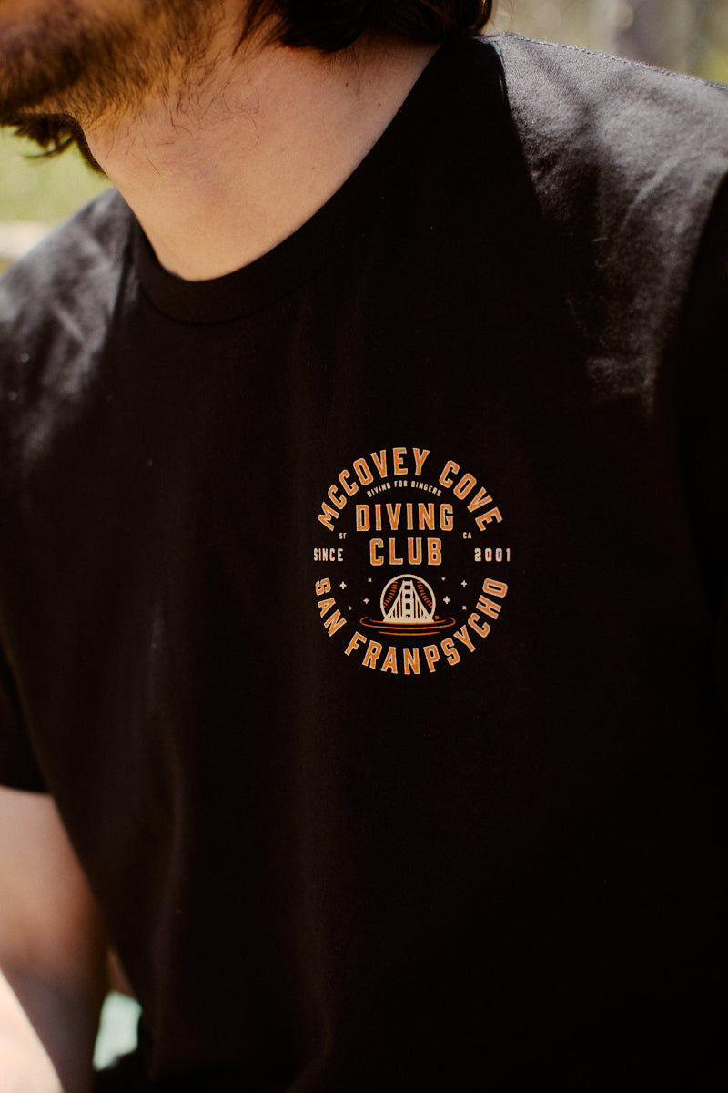 A person dressed in a 100% cotton Giants McCovey Cove Diving Club Tee, showcasing the San Francisco logo in vibrant orange text on a black shirt.