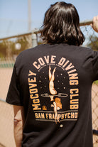 Near a chain-link fence, an individual wears the McCovey Cove Diving Club Tee, a black T-shirt showcasing its distinctive graphic on the back. This 100% cotton tee perfectly complements their collection of Giants apparel, providing both comfort and team spirit.