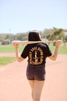 A person in a McCovey Cove Diving Club Tee, made from 100% cotton, holds a bat, standing confidently on a baseball field.