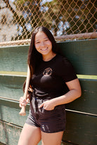 A smiling person, proudly wearing a McCovey Cove Diving Club Tee, holds a baseball bat while leaning against a green wooden fence with a chain-link backdrop.