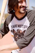 Person smiling in a San Franpsycho Take Meeeow Baseball Tee, holding a bat, standing on a baseball field.