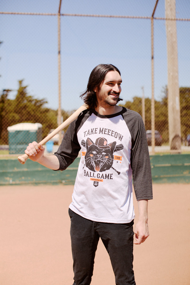 A man smiles with a baseball bat resting on his shoulder, wearing a San Franpsycho "Take Meeeow to the Ball Game" baseball tee on the field.