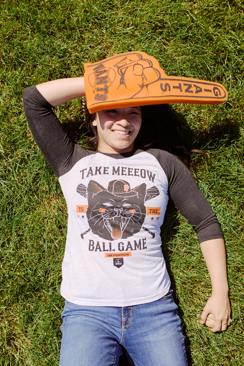 A person relaxes on the grass, sporting a San Franpsycho Take Meeeow Baseball Tee that reads "Take Meow to the Ball Game," and holds a large foam finger.