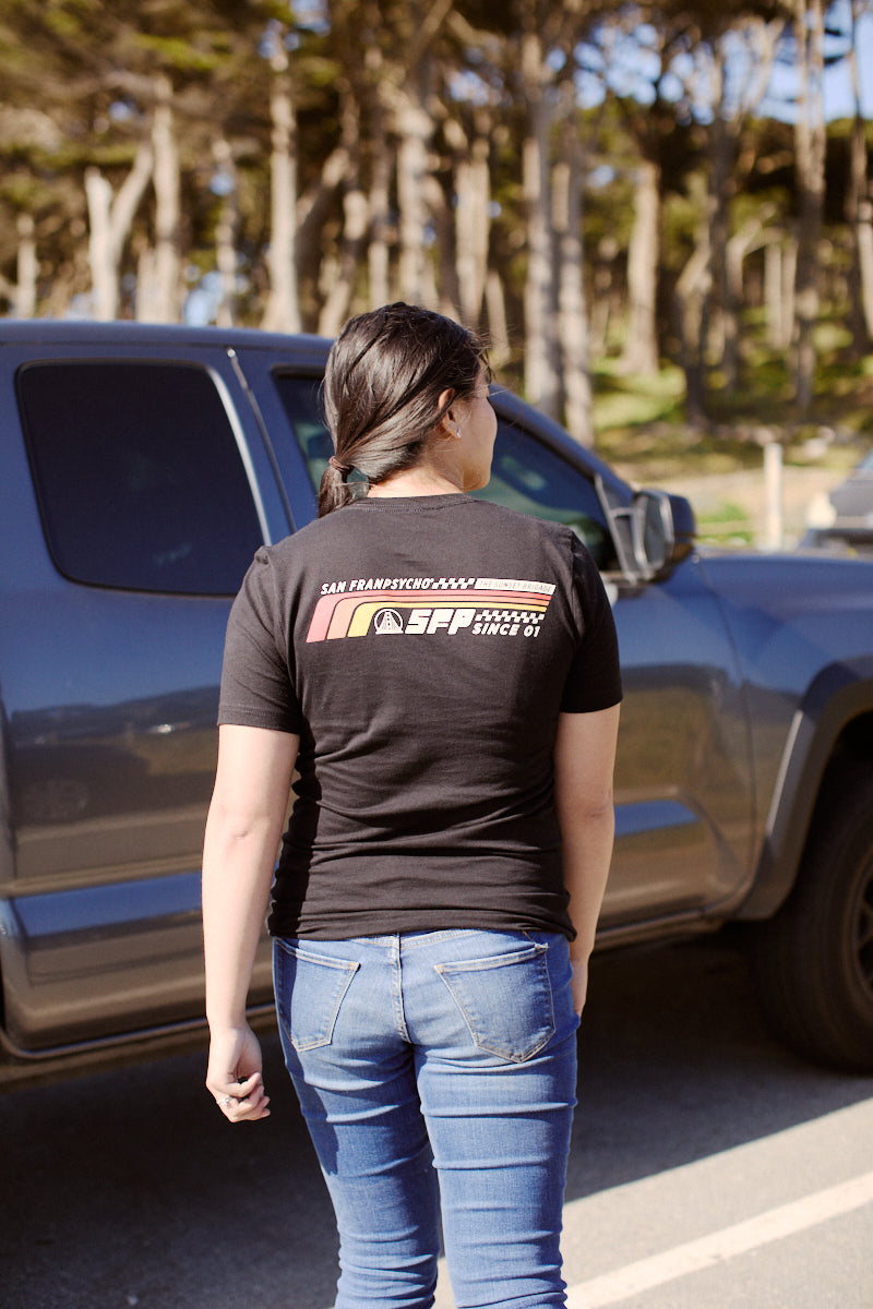 A person wearing a Racing Stripes Tee, made from 100% Airlume cotton, is standing next to a gray truck in a wooded parking area.