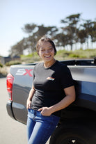 A person grins while resting against a truck, wearing the hand-printed Racing Stripes Tee created from 100% Airlume cotton, with trees softly swaying behind them.