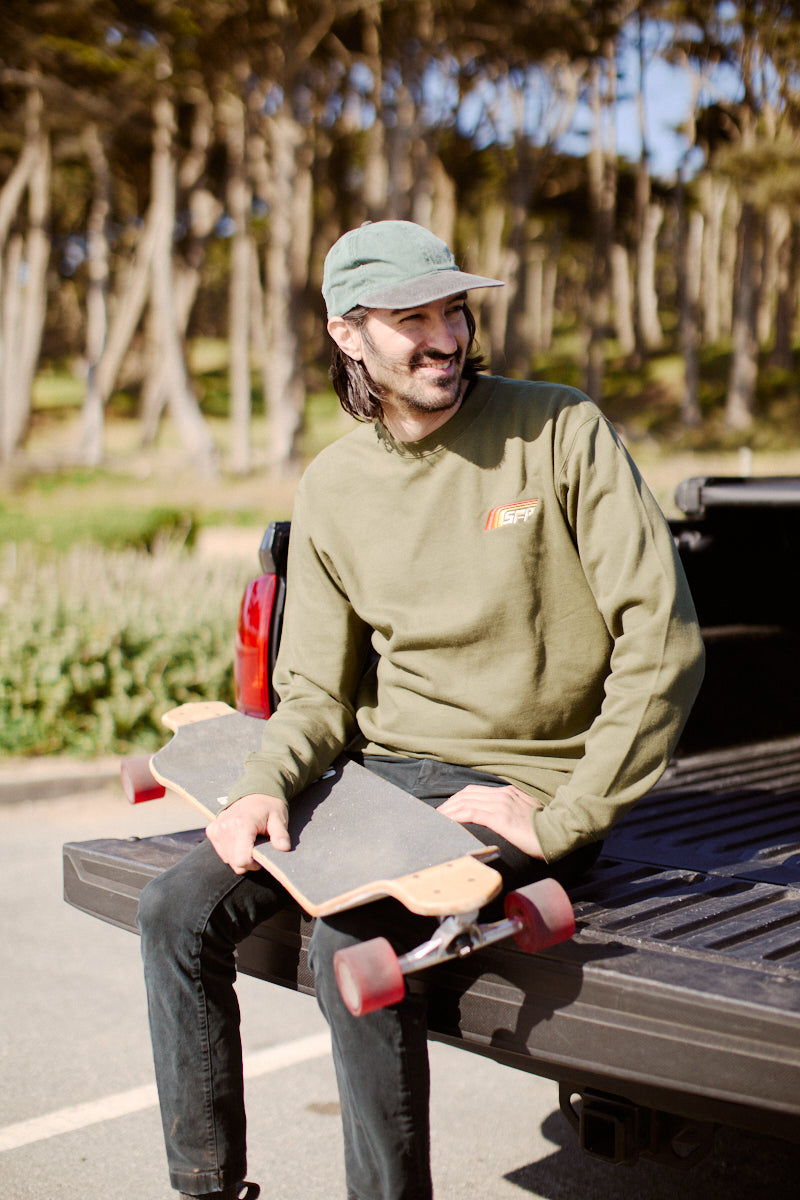 A person sitting on a truck bed holding a skateboard, dressed in the Racing Stripes Crewneck with an 80s-inspired design, surrounded by trees.