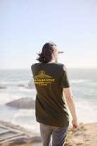 A person with long hair wearing a green Breakers Tee, crafted from soft Airlume cotton by San Franpsycho, gazes over ocean cliffs on a sunny Spring/Summer 2024 day.