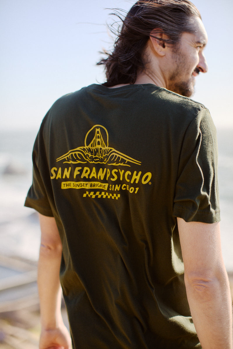 A man with long hair wearing a Breakers Tee by San Franpsycho stands outside by the ocean, savoring the fresh breeze of Spring/Summer 2024. The soft Airlume cotton of his shirt gently billows in the wind, perfectly embodying laid-back coastal style.