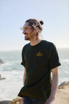 A person with long hair in a bun stands on a beach cliff, wearing a green Breakers Tee crafted from soft cotton, gazing towards the ocean.