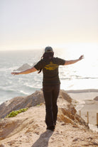 Standing on a cliff edge with arms outstretched, overlooking the ocean and sandy beach, the person is comfortably dressed in the Breakers Tee from our Spring / Summer 2024 collection. Perfect for enjoying breathtaking views, it combines nature's tranquility with contemporary fashion.