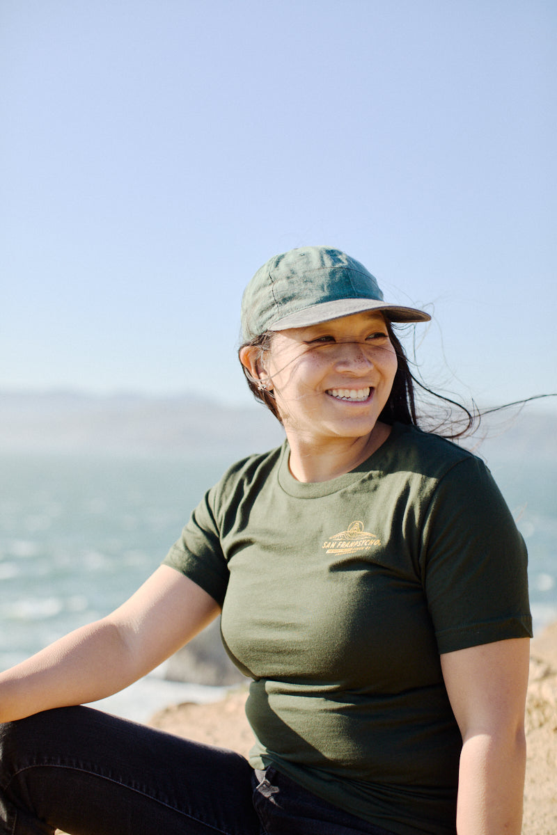 Dressed in a green Breakers Tee from the Spring/Summer 2024 collection, a person enjoys the sunny day while sitting outdoors near the ocean, smiling contentedly.