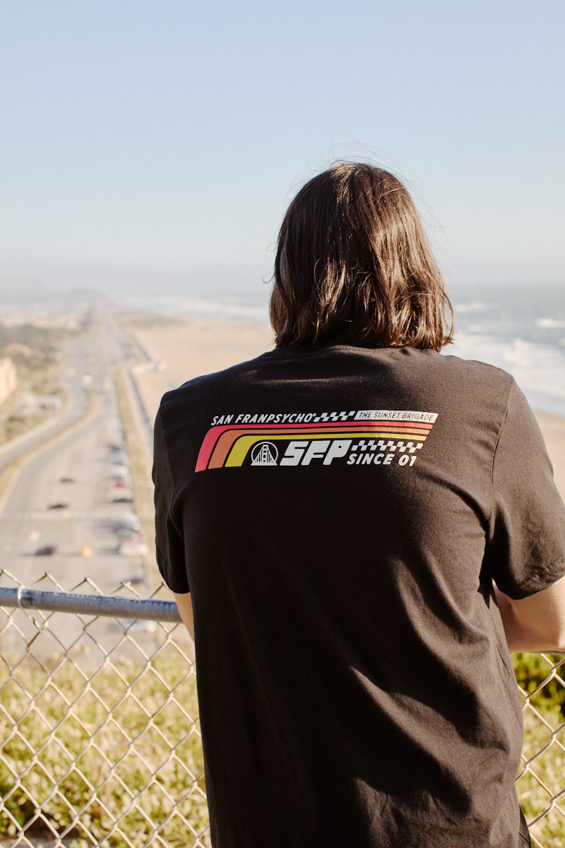 A person with long hair overlooks a coastal road, wearing the Racing Stripes Tee. Crafted from 100% Airlume cotton, this shirt showcases a hand-printed graphic design on the back.