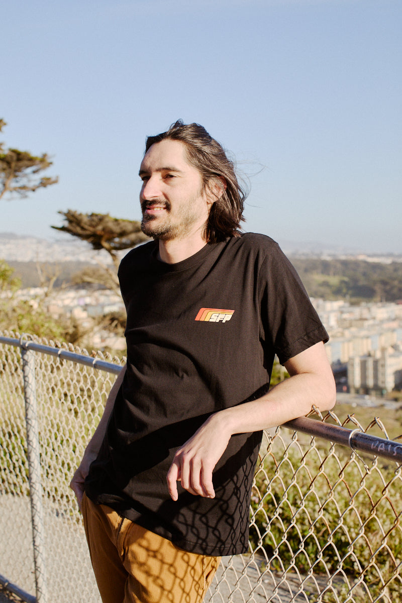 A man with long hair, wearing a Racing Stripes Tee crafted from 100% Airlume cotton, leans on a fence with a cityscape and trees in the background.