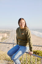 A person sits on a fence, smiling, with a coastline and highway in the background. They're wearing the Racing Stripes Crewneck, its embroidered logo catching the sunlight and adding a touch of nostalgia to the serene scene.