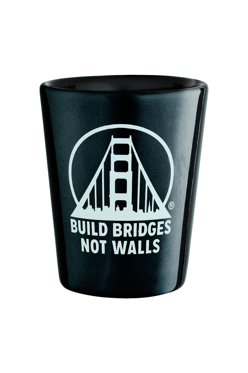 The Build Bridges Not Walls Shot Glass features a black design with a white bridge graphic and the bold San Franpsycho mantra, ideal for those who value connection over division.