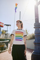 A person beams joyfully, wearing the Rainbow Stripes Tee, standing outdoors as a pride flag waves proudly in the background near the SF LGBT Center.