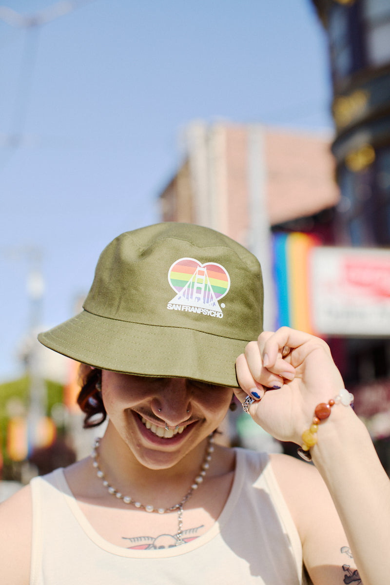 A person is smiling while wearing a Rainbow Logo Bucket Hat, featuring a rainbow logo and the text "San Francisco," near the SF LGBT Center in an urban setting.