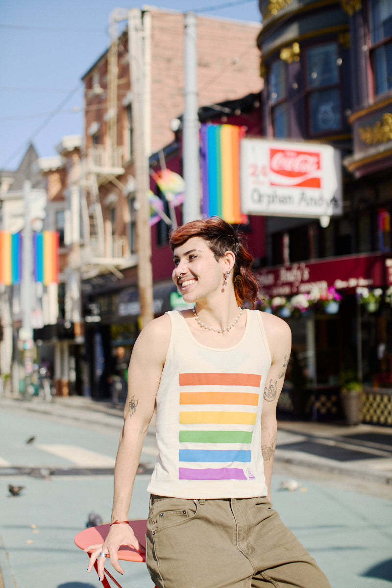 A person beams brightly in a Rainbow Stripes Tank Top, holding their skateboard on a lively city street adorned with rainbow flags. The vibrant scene echoes the spirit of unity and pride, reminiscent of the inclusive energy found near the SF LGBT Center.