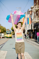 A person joyfully waves a transgender pride flag on a sunny street, their Rainbow Stripes Tank Top catching the light. In the background, rainbow flags flutter and a tram glides past, capturing the vibrant spirit near the SF LGBT Center.