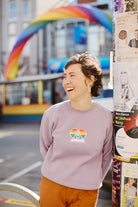 A smiling person in a Rainbow Heart Crewneck stands by a pole adorned with posters, with the SF LGBT Center's rainbow arch in the background.