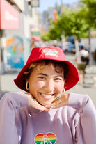 A person smiling, wearing a Rainbow Logo Bucket Hat and a sweatshirt with a rainbow logo featuring a bridge design, stands against a backdrop of trees and buildings reminiscent of the SF LGBT Center.