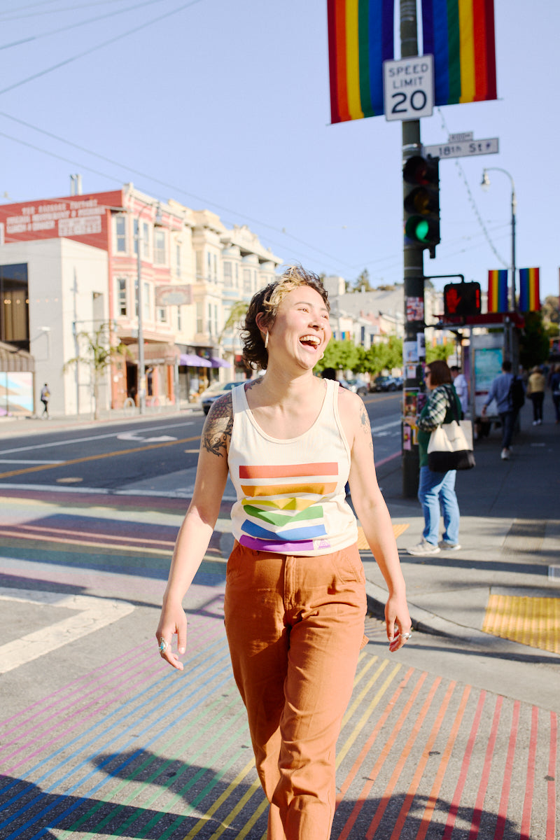 A person, smiling and embracing the vibrant energy of the city, walks across a colorful crosswalk adorned with rainbow flags. They're wearing a Rainbow Stripes Tank Top, perfectly matching the lively urban setting in front of the SF LGBT Center.