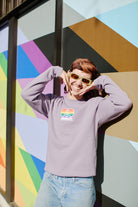 Wearing a Rainbow Heart Crewneck and sunglasses, a person poses happily against a colorful geometric mural.