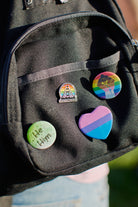 Close-up of a backpack adorned with the Rainbow Logo Pin and various colorful enamel pins and badges, including He/Him and a heart, showcasing PRIDE in every detail.