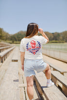 Dressed in an Eternal Flame Tee and denim shorts, a person stands on outdoor bleachers beneath a clear blue sky, epitomizing the vibrant spirit of San Francisco during the Olympic season.