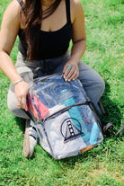 A person sits on the grass with a Clear Backpack w/ SFP Logo, brimming with colorful items. They are dressed in a black top and gray jeans, ready to soak up the festival vibes around them.