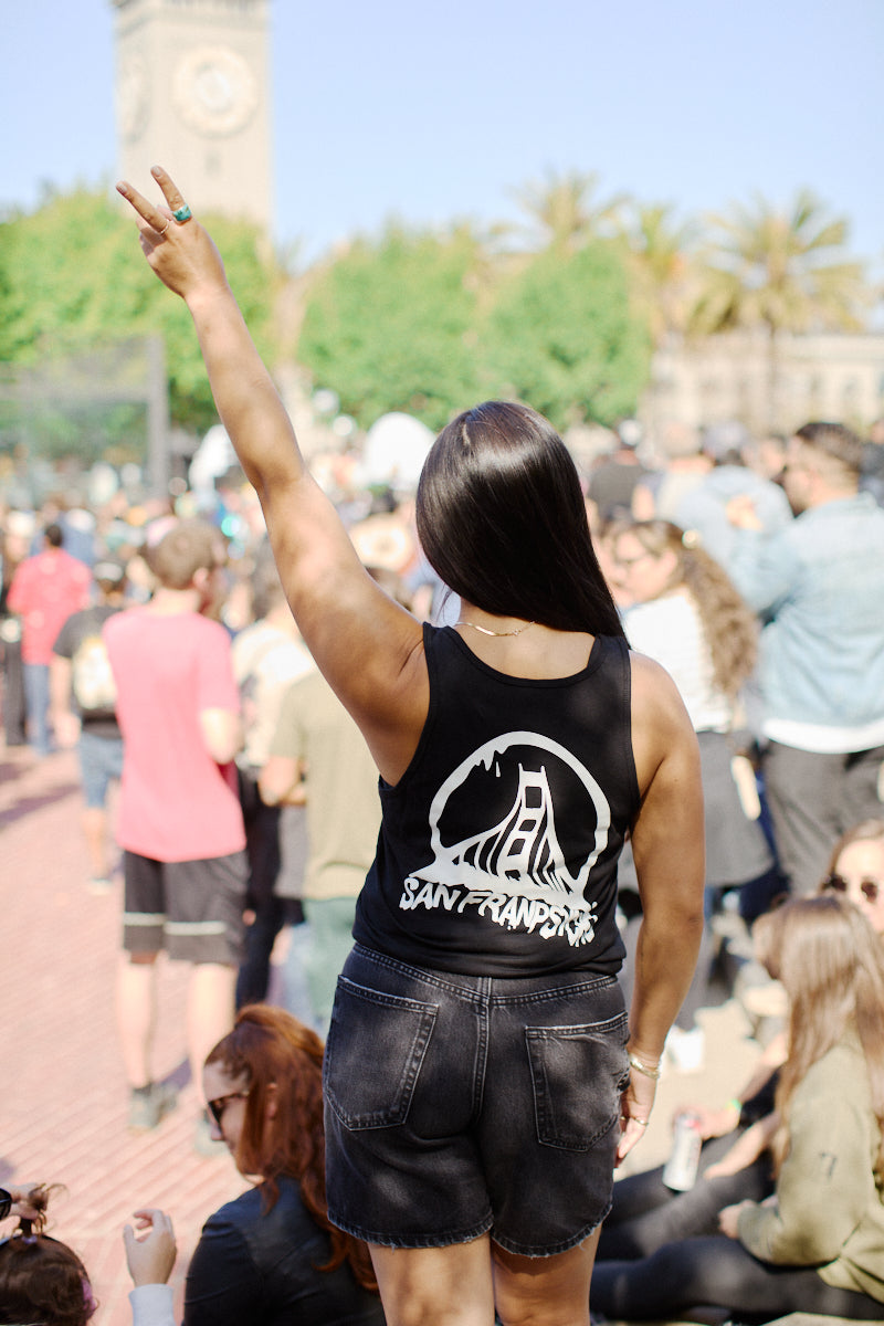 Wearing the fashionable Acid Logo Tank Top, a person raises their hand in a peace sign at an outdoor event surrounded by a lively crowd and the iconic San Francisco clock tower.