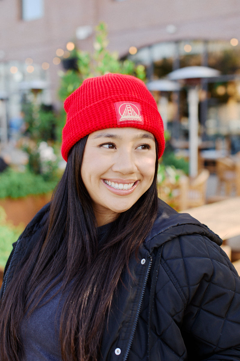 A smiling person wearing a vibrant Red Waffle Beanie featuring the Red/Gold Logo and a sleek black jacket enjoys the outdoor setting, embodying the chill vibes of San Franpsycho.