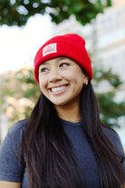 A woman wearing a vibrant Red Waffle Beanie with a natural/red logo patch and a gray San Franpsycho t-shirt smiles outdoors, framed by lush trees and towering buildings in the background.