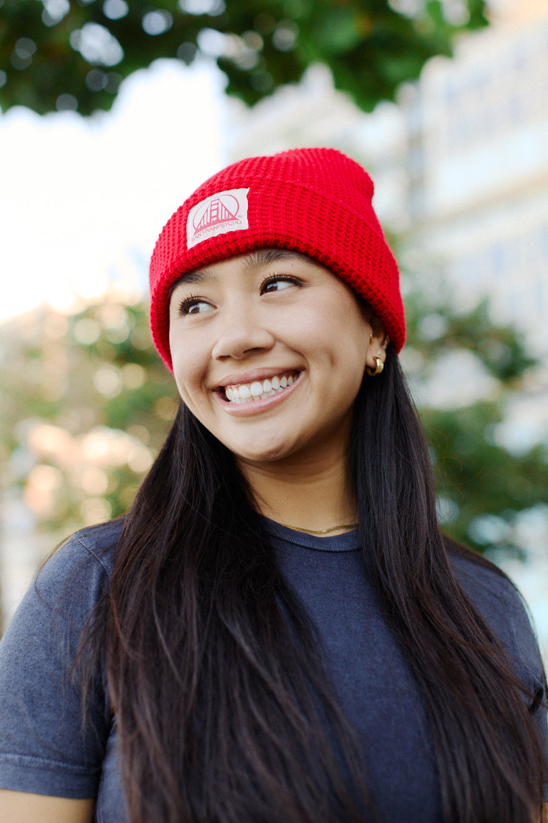 A woman wearing a vibrant Red Waffle Beanie with a natural/red logo patch and a gray San Franpsycho t-shirt smiles outdoors, framed by lush trees and towering buildings in the background.