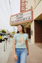 A woman wearing a Womens Cat House Tee - Sage and jeans strolls past a theater marquee. Her T-shirt, hand-printed in San Francisco from 100% ring-spun cotton, showcases a unique graphic design, making it stand out.