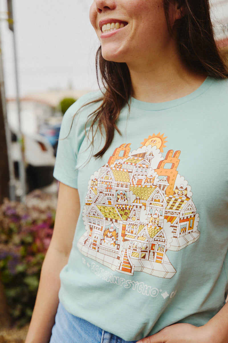 A person beams while wearing the Women's Cat House Tee in light blue, adorned with a vibrant illustrated cityscape that echoes the artistic flair of Ferris Plock. The design beautifully enhances the tailored cut, making it an artistic addition to any wardrobe.