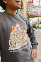 A person wearing the Cat House Crewneck, a gray unisex sweatshirt featuring a colorful vintage character graphic of San Francisco houses, is standing outdoors.