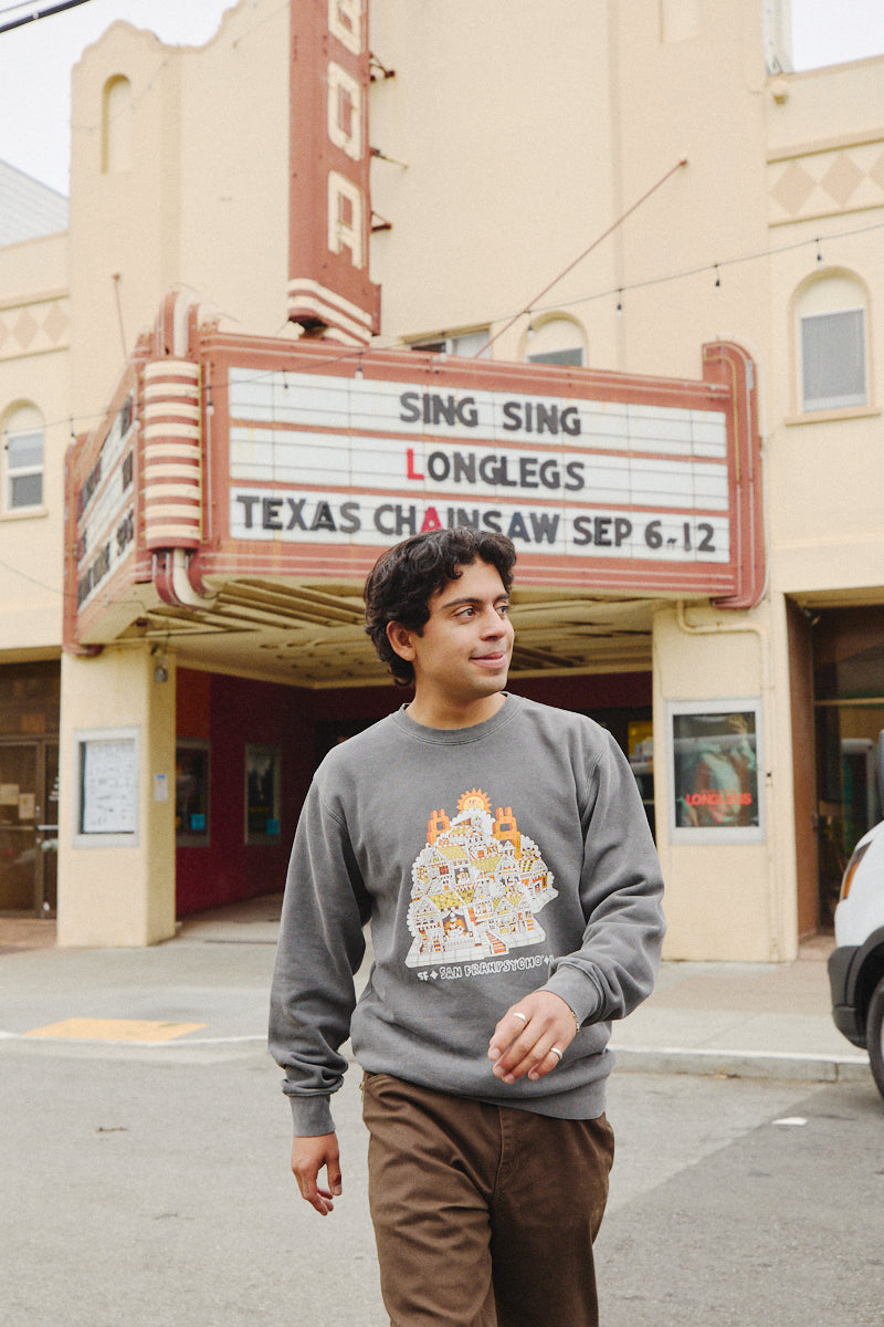 A person strolls in front of a vintage theater, its marquee showcasing movie titles, while sporting the Cat House Crewneck that perfectly complements the theater's vintage character.