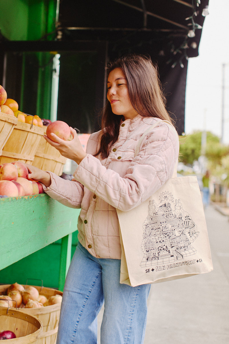 A person in a pink jacket selects apples from a market stand, carrying a Cat House Tote adorned with an urban design by Ferris Plock.